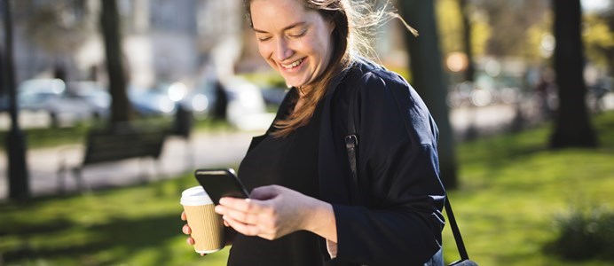 ung kvinna på promenad utomhus, hon håller sin mobil i ena handen och i den andra håller hon en kaffekopp