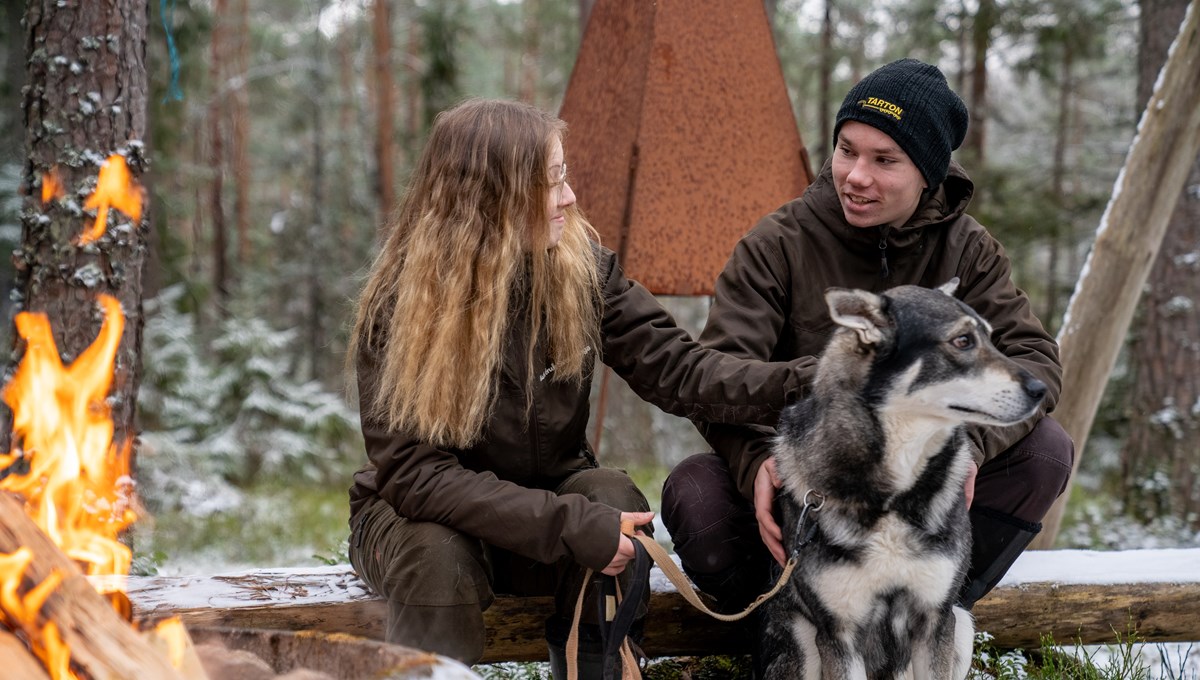 Två elever sitter tillsammans med en hund framför brasa i skogen