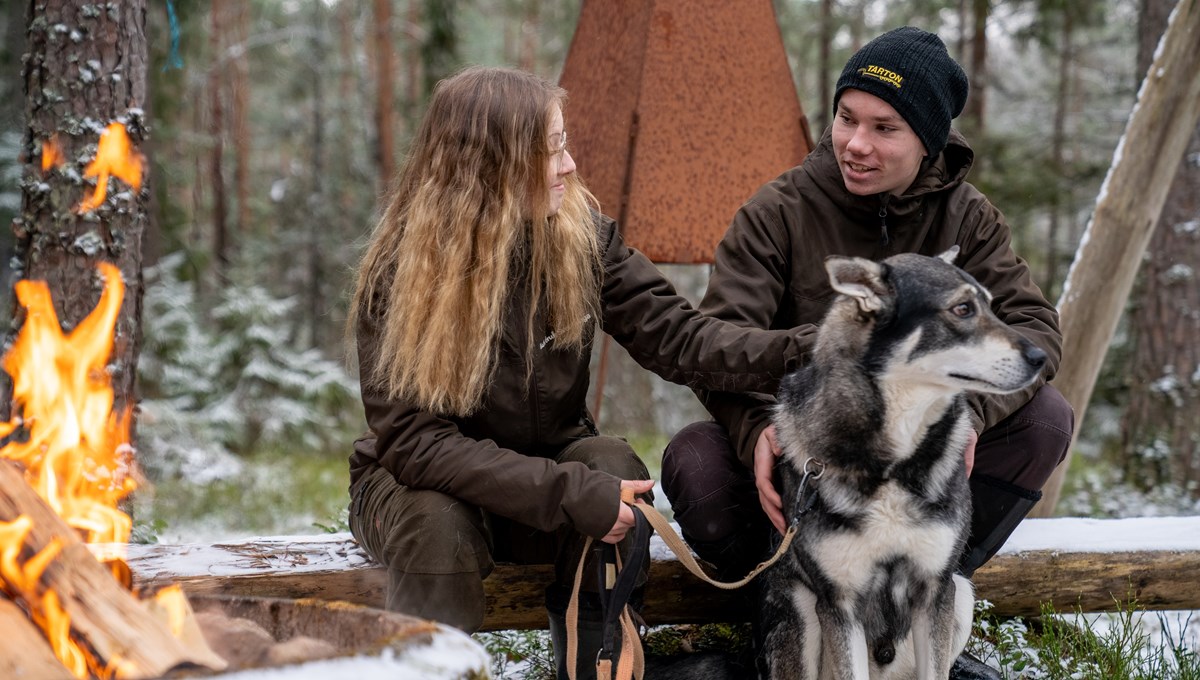 Två friluftsklädda elever sitter framför brasa i skogen med en hund