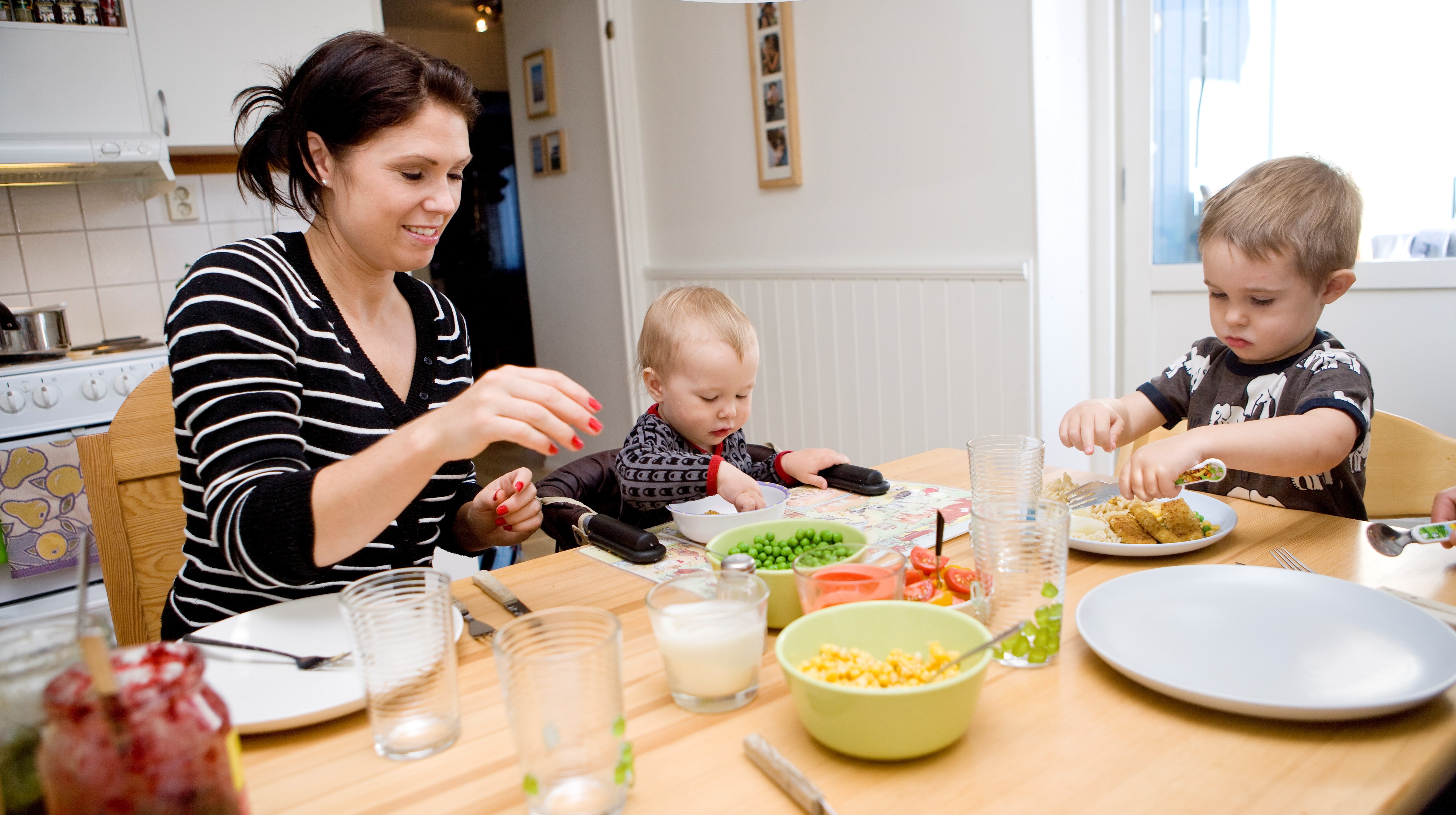 En mamma och två barn sitter vid ett köksbord och äter. På bordet grönsaker i skålar.