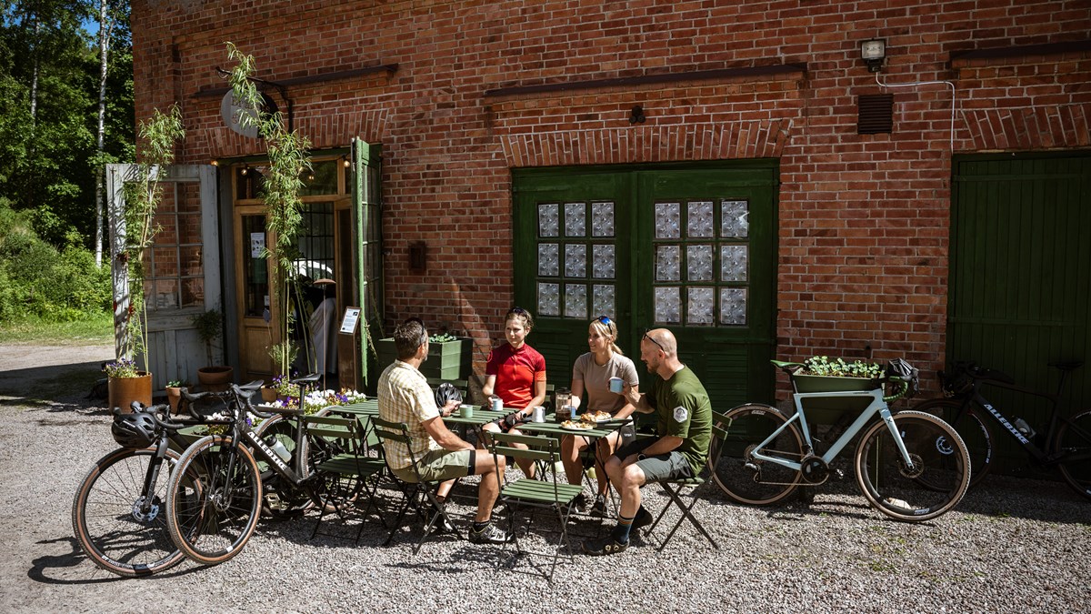 Fyra personer fikar utomhus med sina cyklar uppställda i gruset, framför en äldre röd tegelbyggnad