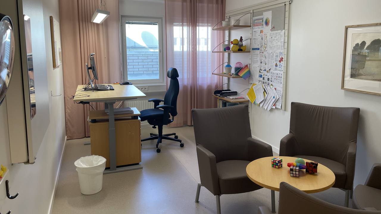 Behandlingsrum med grå fåtöljer vid ett runt litet bord. Längre bort i rummet finns ett skrivbord med en dator och en kontorsstol.