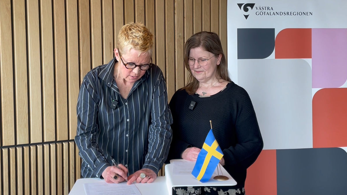 Två kvinnor står vid en pulpet och den ena signerar ett dokument. 