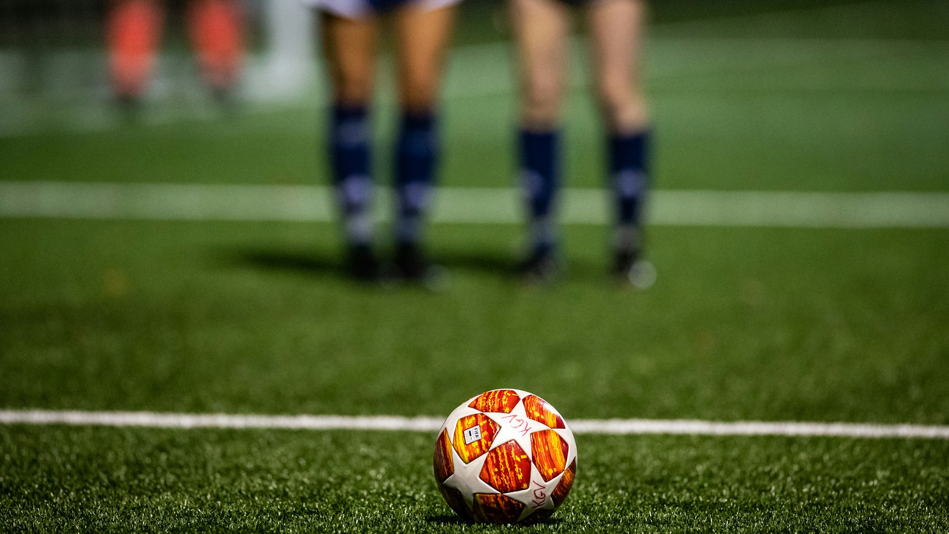 En fotboll ligger på en konstgräsplan. I bakgrunden står två fotbollstjejer, de är i oskärpa och man ser endast deras ben med blå fotbollstrumpor till knäna och shorts.