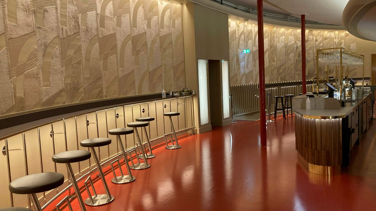 Den nyrenoverade foajébaren på Stadsteatern i Göteborg. en böjd fondvägg klädd med den gyllene tapeten med geometriskt mönster. Utmed väggen löper en väggfast smal borsyta med barstolar placerade med jämna intervall. Till höger i rummet en bardisk klädd i trä. Golvet är rött och högblankt.