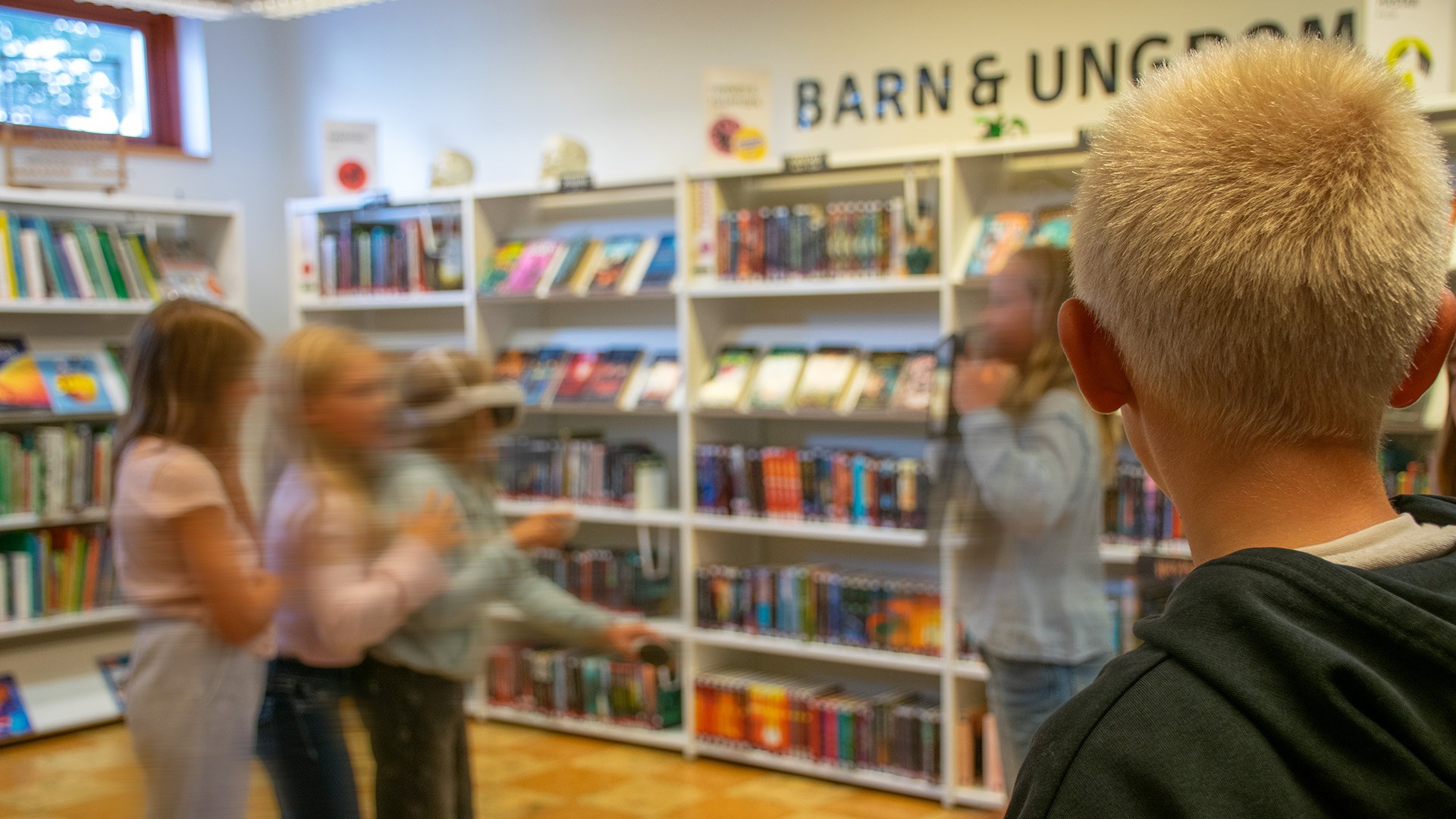 En pojke sitter i förgrunden, hans blick är vänd mot tre flickor, varav en har ett VR-headset på sig. Flickorna är oskarpa på bilden, pojkens bakhuvud är skarpt. Runt om i rummet står fyllda vita bokhyllor. På väggen står texten "Barn och ungdom". 