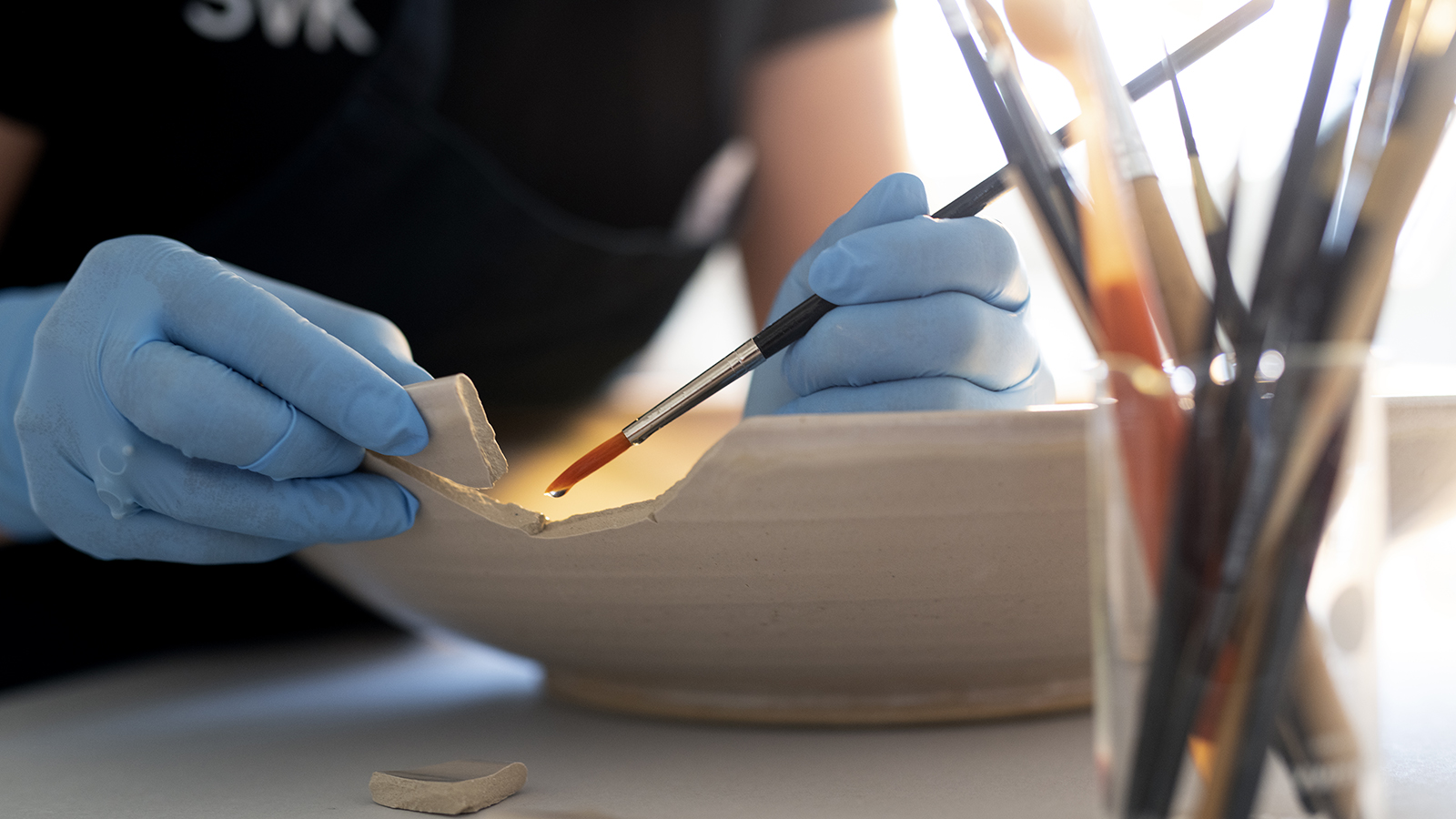 En person med blå latexhandskar limmar försiktigt fast en skärva på ett stort keramikfat.