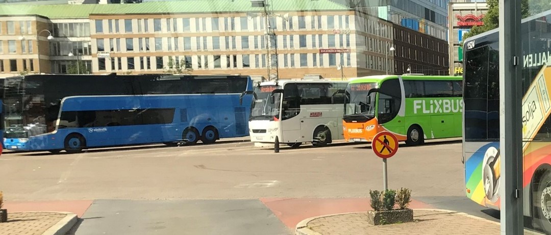 Fotografi genom glasrutan från Nils Ericson-terminalen i Göteborg. Utanför terminalen står parkerade bussar från olika bussföretag. Det är soligt sommarväder och köpcentret Nordstan syns i bakgrunden.