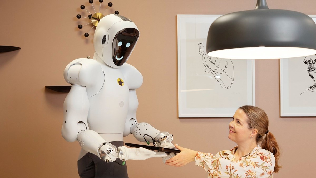 Vit, humaniod robot med runda former räcker laptop till sittande kvinna. 