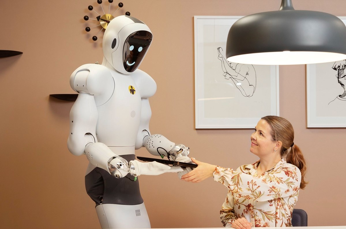 Vit, humaniod robot med runda former räcker en laptop till sittande kvinna. 