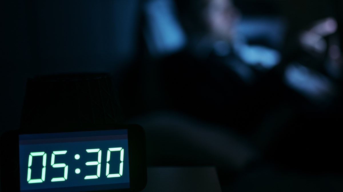 Bilden visar en digital klocka i ett mörkt rum. Klockan visar 05:30. I bakgrunden syns konturerna av en person som sitter framför en skärm.