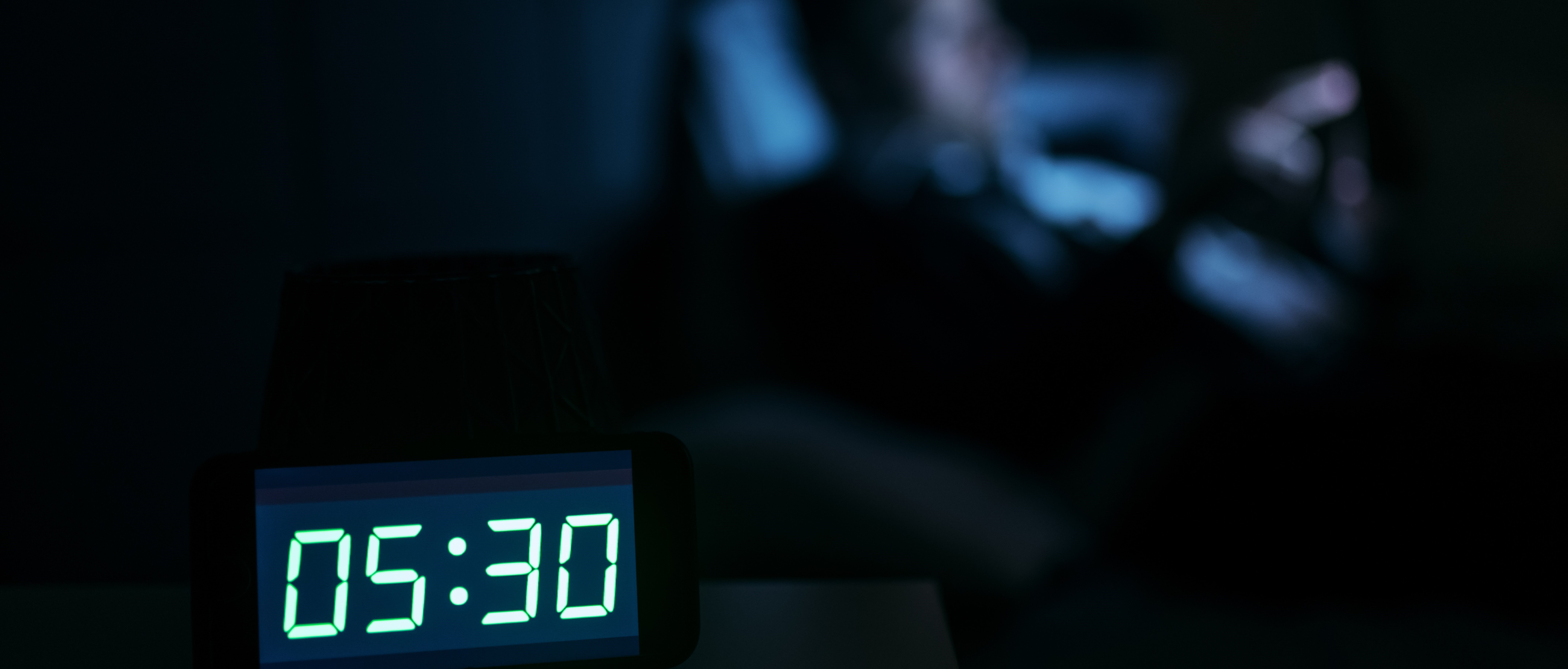 Bilden visar en digital klocka i ett mörkt rum. Klockan visar 05:30. I bakgrunden syns konturerna av en person som sitter framför en skärm.