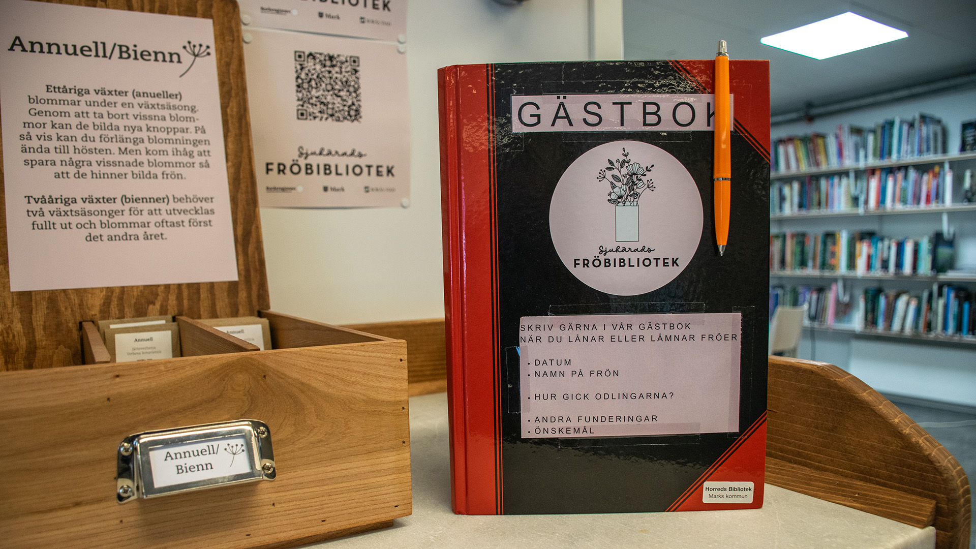 En gästbok med texten Fröbibliotek står på en bänk. På boken står instruktioner till hur man lånar fröer ut fröbiblioteket, Bokhyllor fyllda med böcker finns i bakgrunden. Bredvid boken står en trälåda med texten Annuell Bienn.