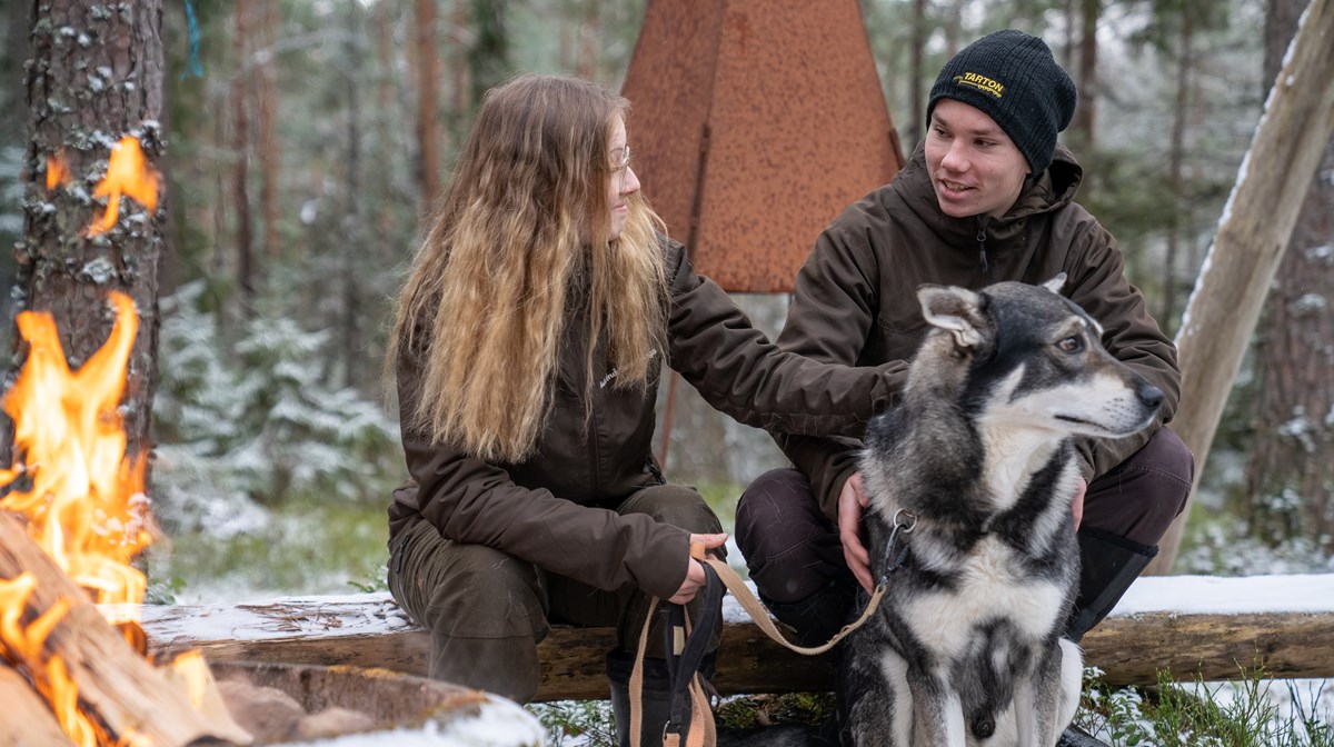 Två elever sitter i skogen framför brasa med hund