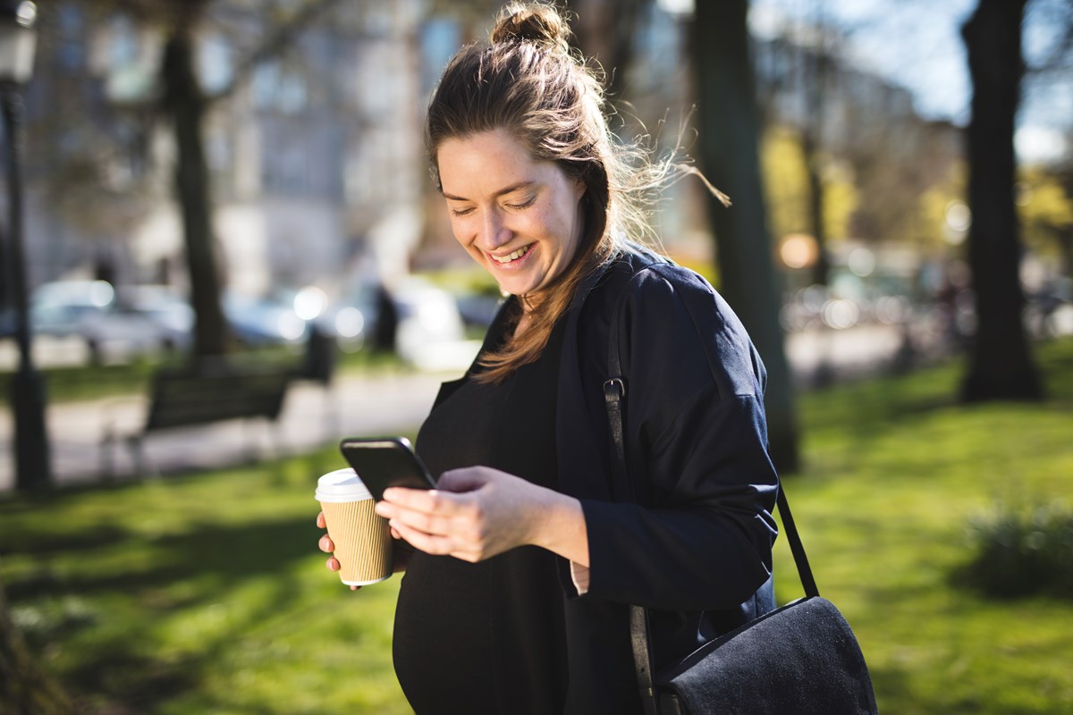 Gravid kvinna vandrar i en park och tittar på sin mobiltelefon.