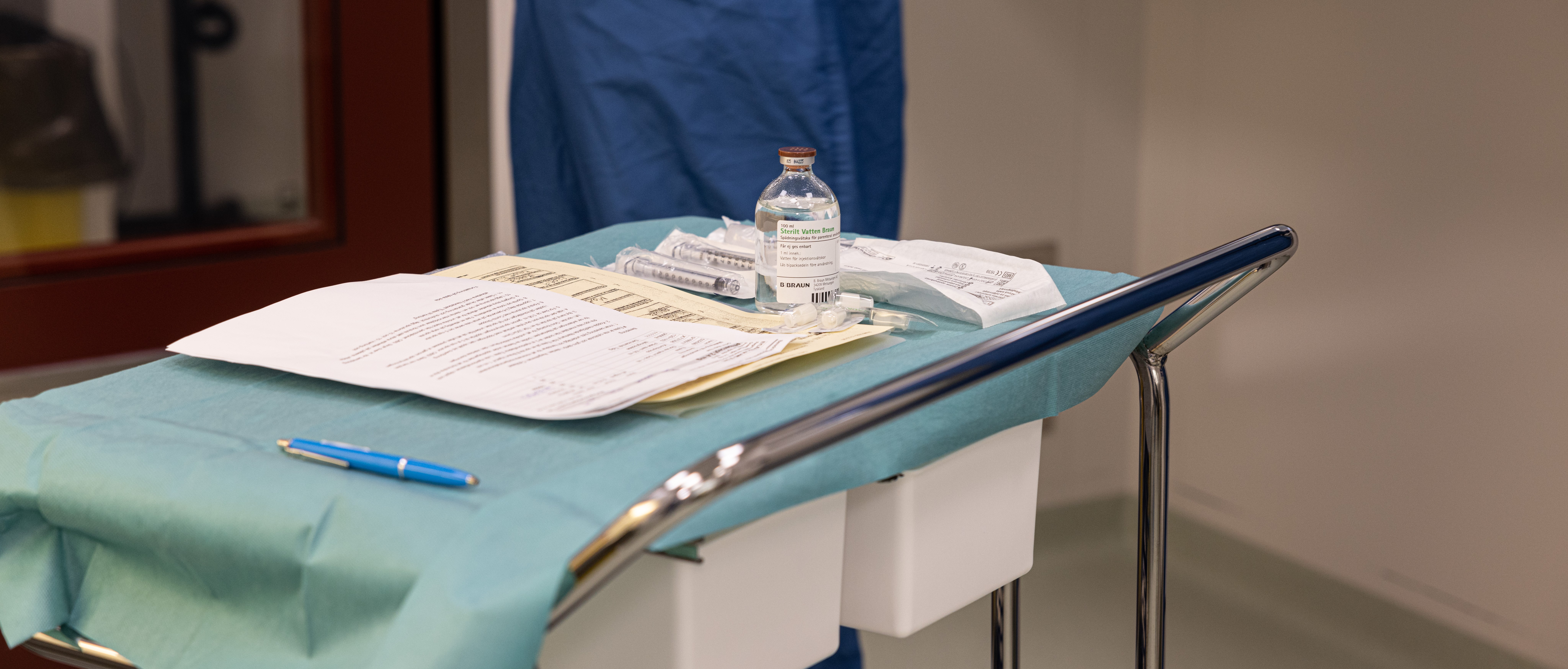  En stålvagn på hjul står i ett rum på ett sjukhus. Vagnen är täckt med blått papper, på vagnen ligger det A4 papper med text, en bläckpenna, en genomskinlig glasflaska med etikett och bredvid ligger det sprutor som är inplastade. I bakgrunden syns en person i blå läkarrock.