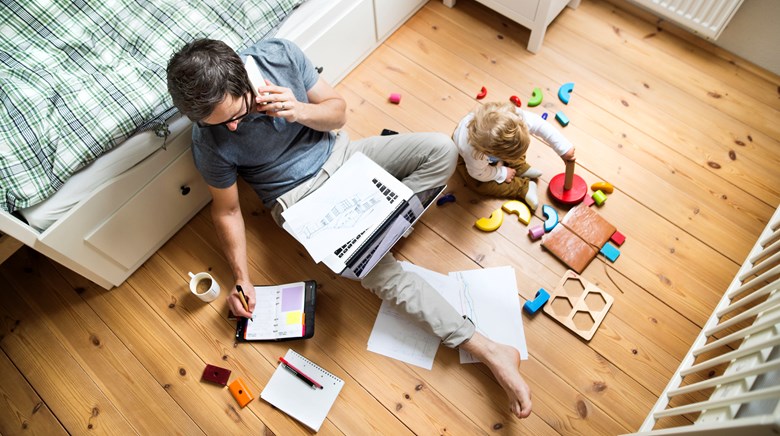 En pappa arbetar hemifrån sittandes på golvet med sittbarn bredvid som leker med klossar
