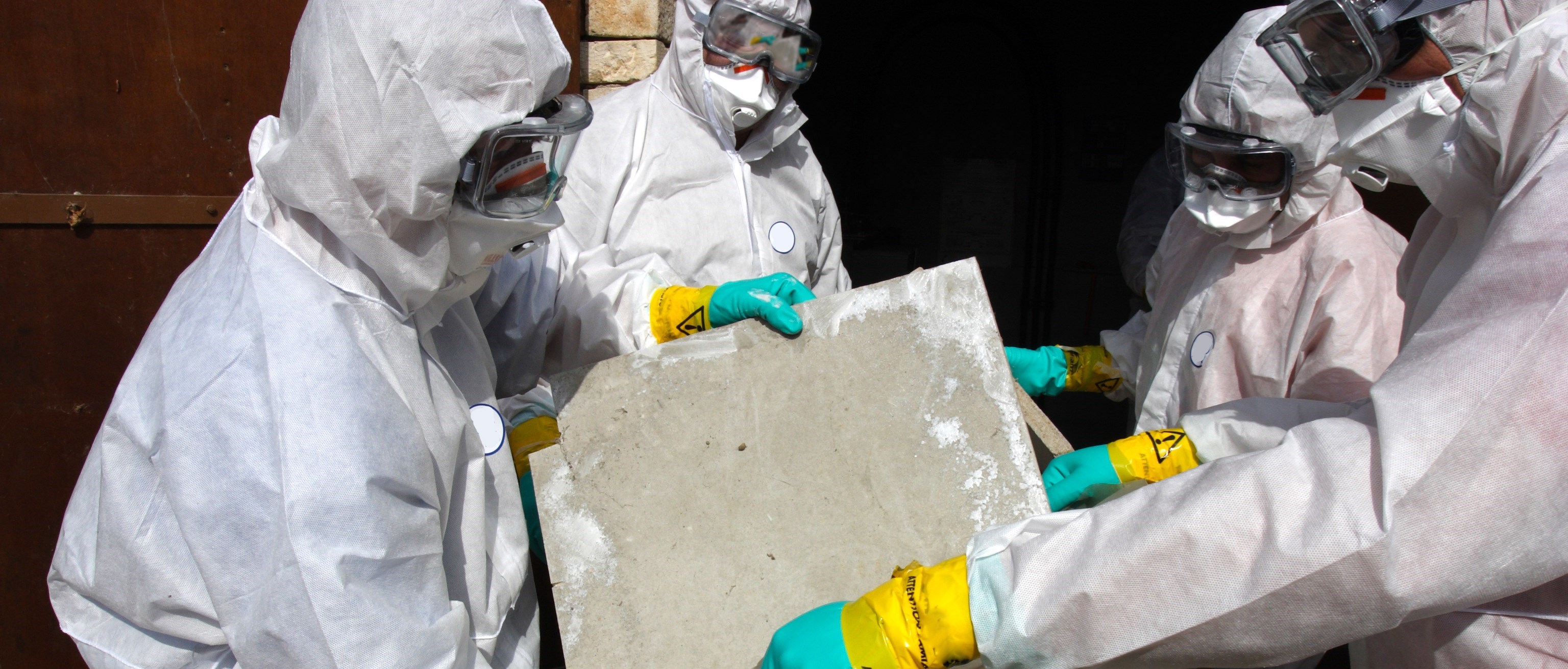 Halvbild som visar fyra personer i heltäckande skyddskläder, munskydd och skyddsglasögon. De håller en skiva med asbest som de lägger i en saneringspåse. Personerna står bakom en avspärrning.