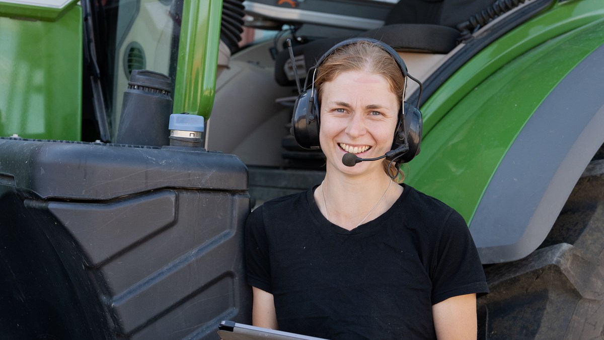 Kvinnlig servicetekniker med hörselkåpor står framför traktor och håller i en laptop
