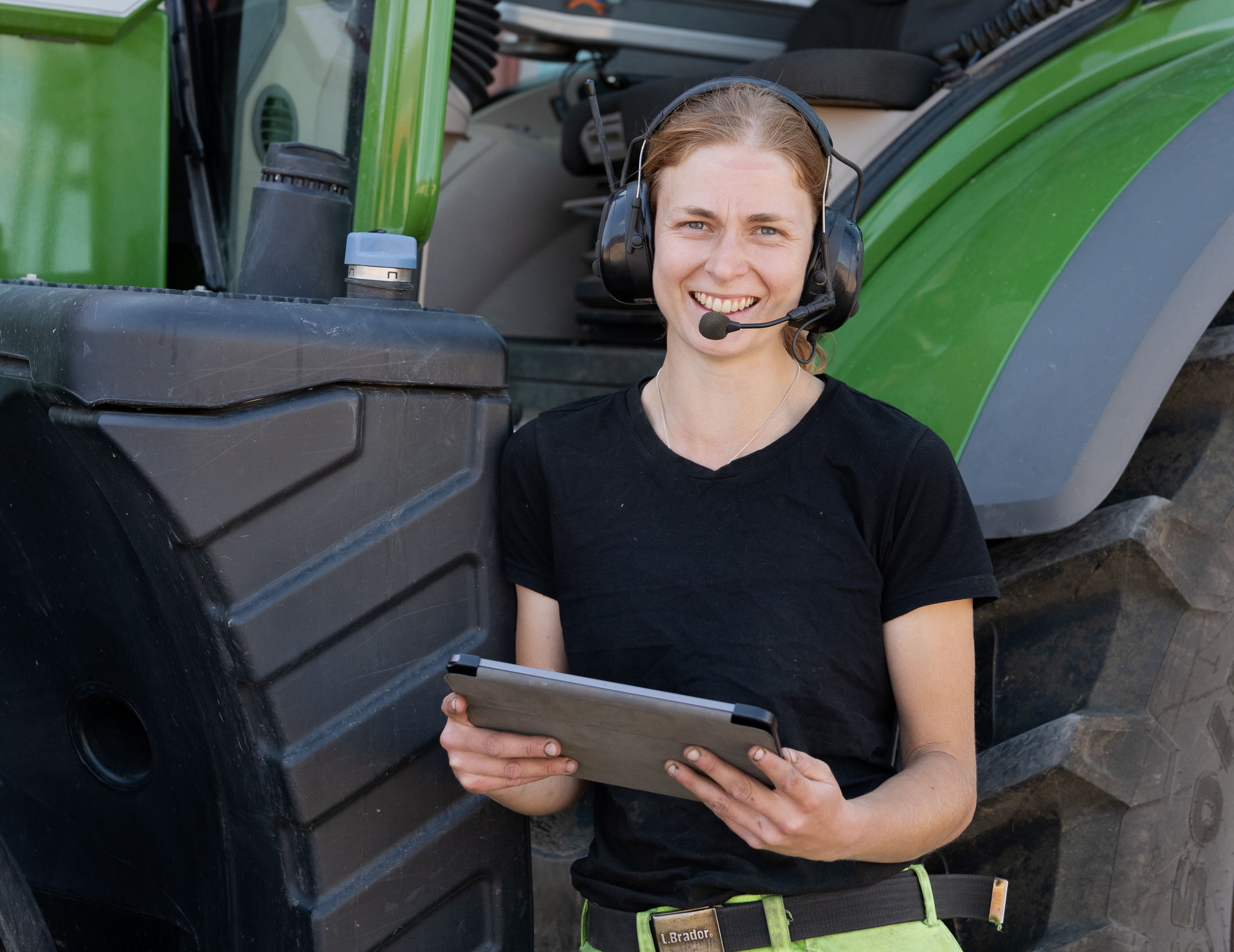 Kvinnlig servicetekniker med hörselkåpor står framför traktor och håller i en laptop