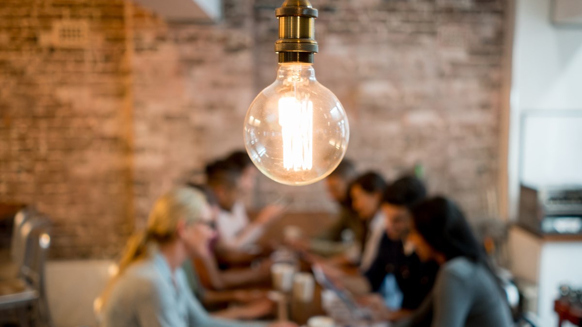 Bild med en lysande glödlampa i förgrunden. Bakgrunden är suddig och visar en grupp människor som sitter och arbetar tillsammans runt ett bord.