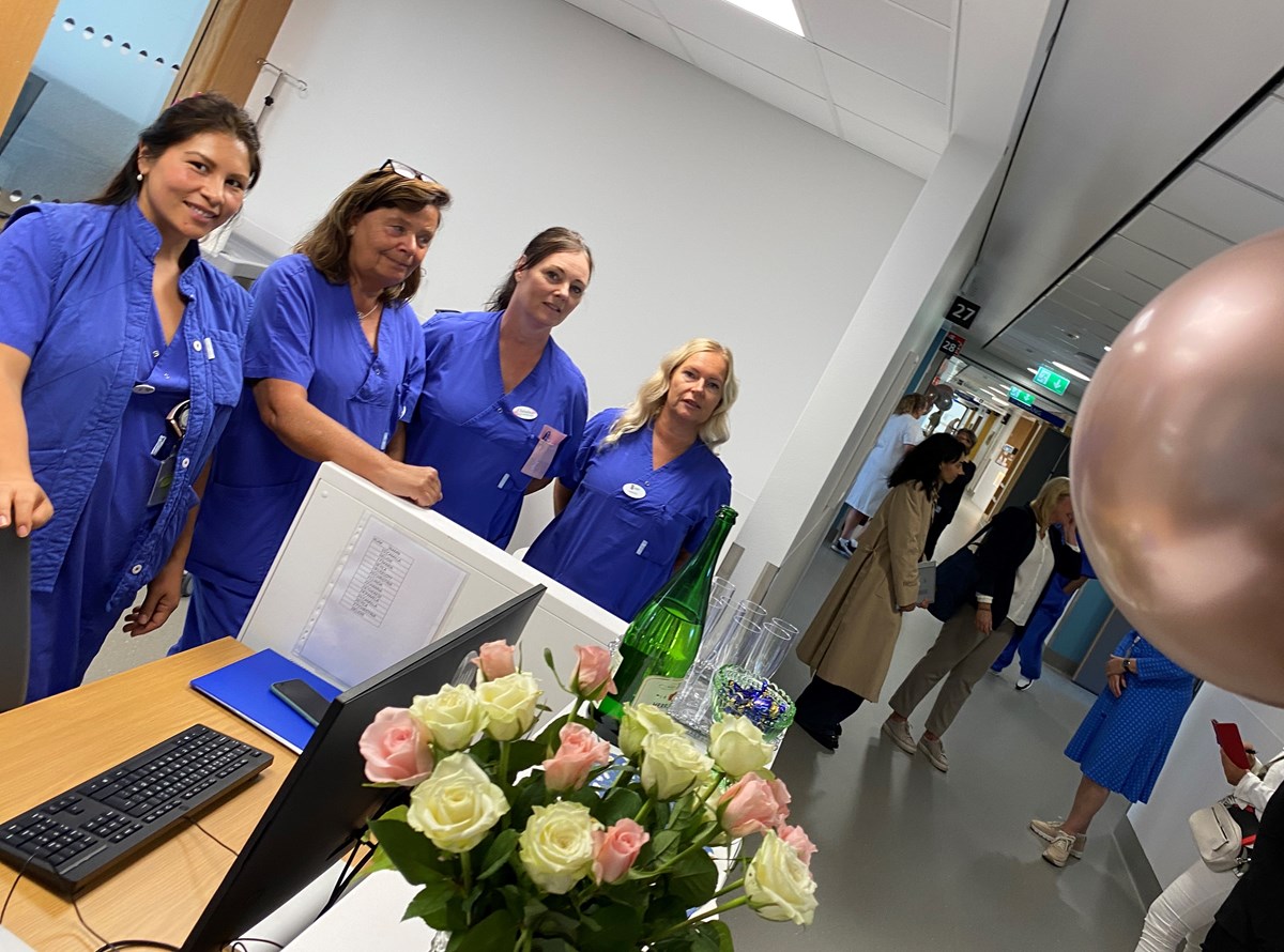 Balong och blommor i förgrunden. Fyra blåklädda sjuk- och undersköterskor. Minglande personer i korridor i bakgrunden.