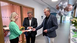 Enhetschef i grön blus får ta emot blomma och kort från Sjukhusen i västers styrelseordförande. Med på bilden också kommunstyrelsens ordförande. Ballonger hänger i taket