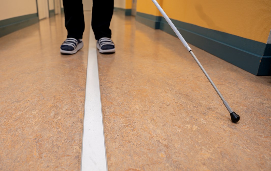 Två fötter på varsin sida av ett ledstråk i en korridor. Till höger finns en vit käpp.