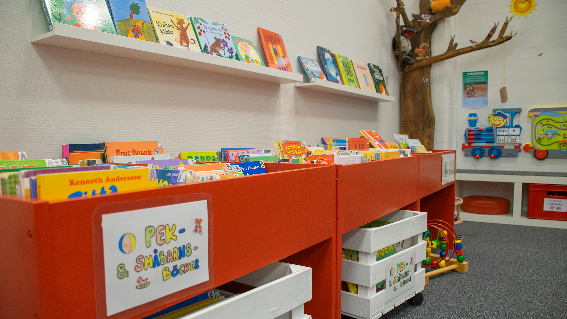 Röda låga boktråg med skylten Pek- & småbarnsböcker. På den vita väggen sitter lister där fler böcker står uppställda. Vita backar med hjul står på golvet, även de fyllda med böcker. Mattan är grå.