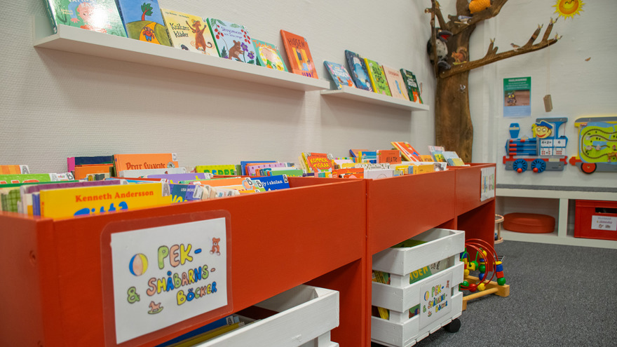 Röda låga boktråg med skylten Pek- & småbarnsböcker. På den vita väggen sitter lister där fler böcker står uppställda. Vita backar med hjul står på golvet, även de fyllda med böcker. Mattan är grå.