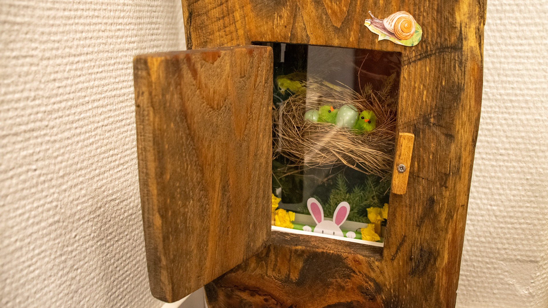 I ett stiliserat träd finns en lucka. Luckan är öppen och innanför finns ett litet fack med ett fågelbo med små gröna småfågelungar i. 