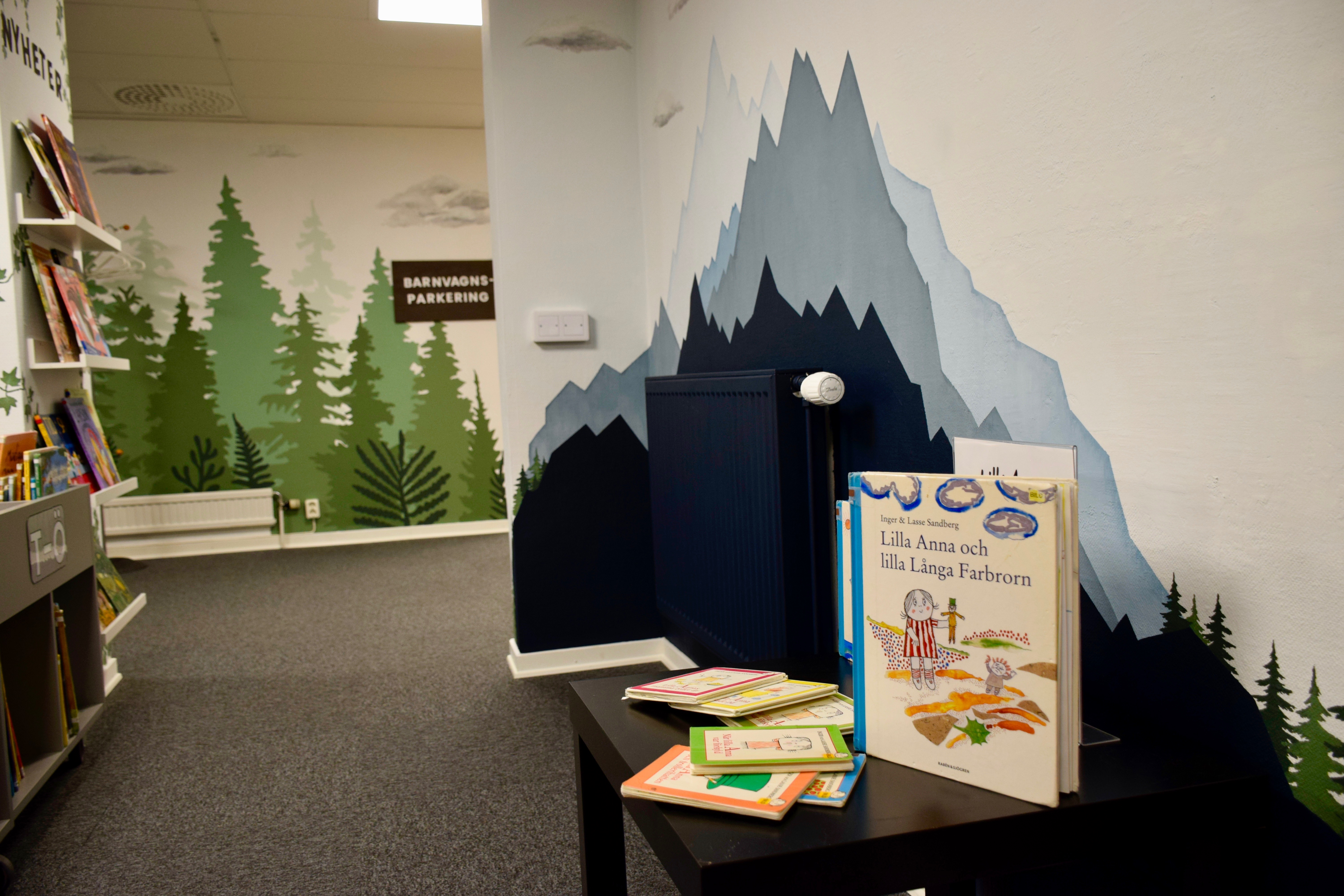 En vägg är målad med spetsiga berg i olika blå kulörer. Framför väggen står ett bord med uppställda böcker. 