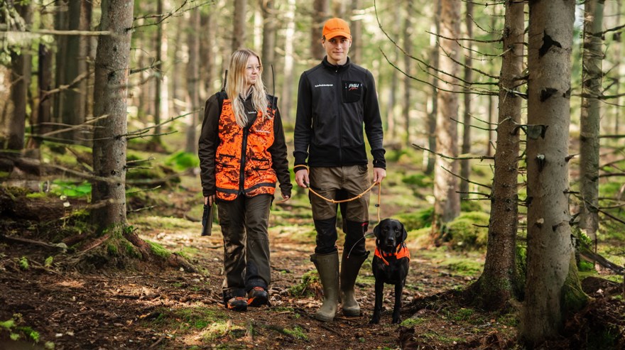 Två elever i jaktkläder promenerar i skogen med hund