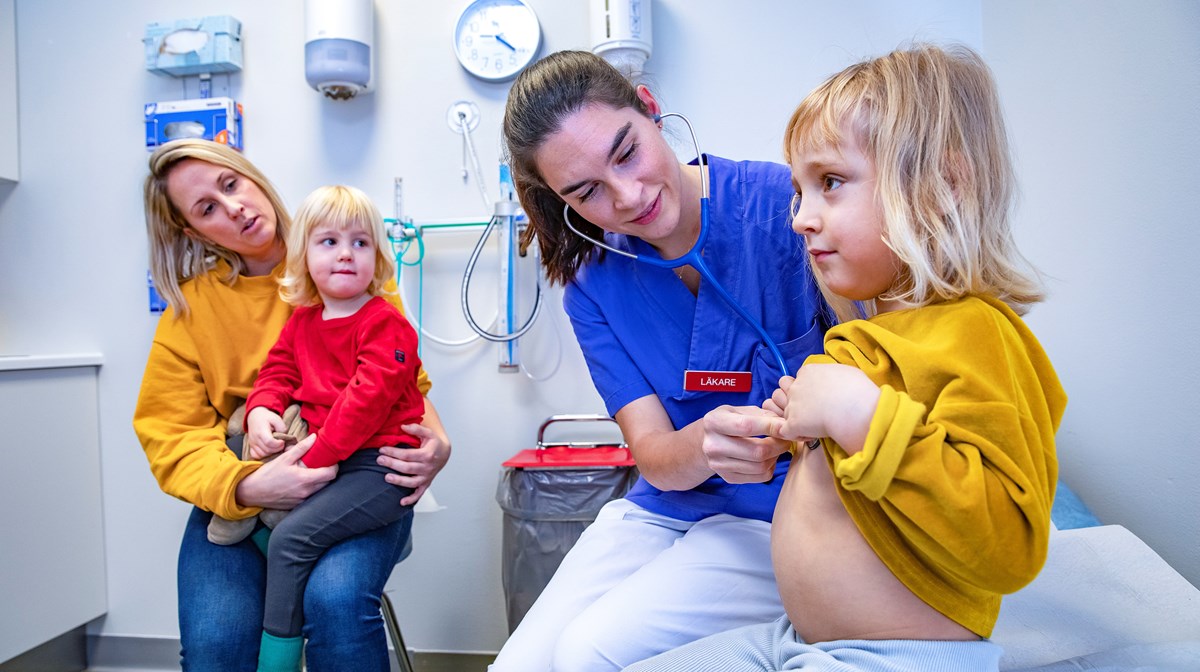 En kvinnlig ung läkare med lyssnar på en flickas hjärta i ett undersökningsrum. Med i rummet finns flickans mamma med ett barn i knät.