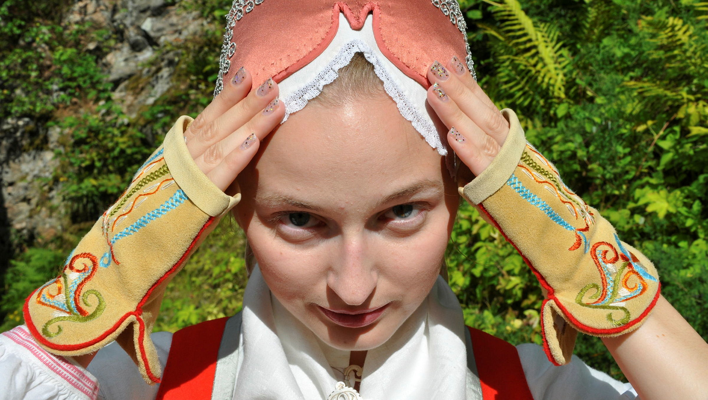 Dräktklädd kvinna som håller sina händer med broderade halvvantar mot stycket och bindmössan på sitt huvud. 