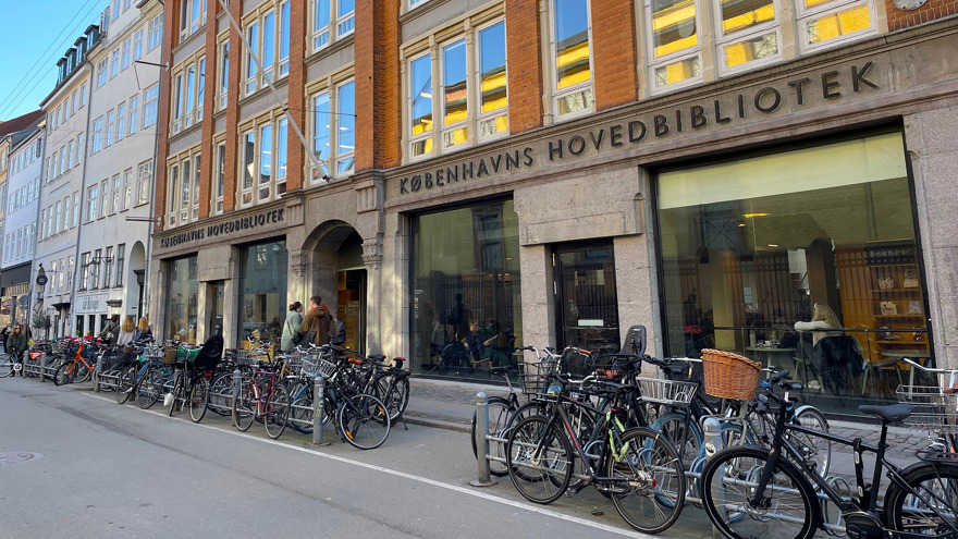 En gata i Köpenhamn. En byggnad med stora skyltfönster mot gatan, utanför står många cyklar parkerade. Ovanför ingången står det med stora bokstäver KÖPENHAMNS HUVUDBIBLIOTEK på danska.