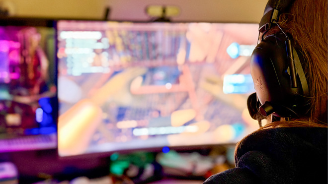 En person sitter framför en dator. Bilden är tagen bakifrån och man ser att personen bär hörlurar. På skärmen finns en spelplan av något slag, skärmen är oskarp och färgglad.