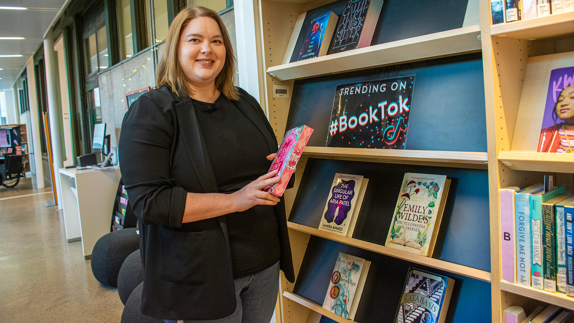 Erika Greve är i 30-årsåldern och har axellångt ljusbrunt hår. Hon står vid en bokhylla på biblioteket som har skylten TRENDING ON #BOOKTOK. Hon håller i en bok och tittar in i kameran. Erika är klädd i jeans och en svart kavaj med en svart topp.