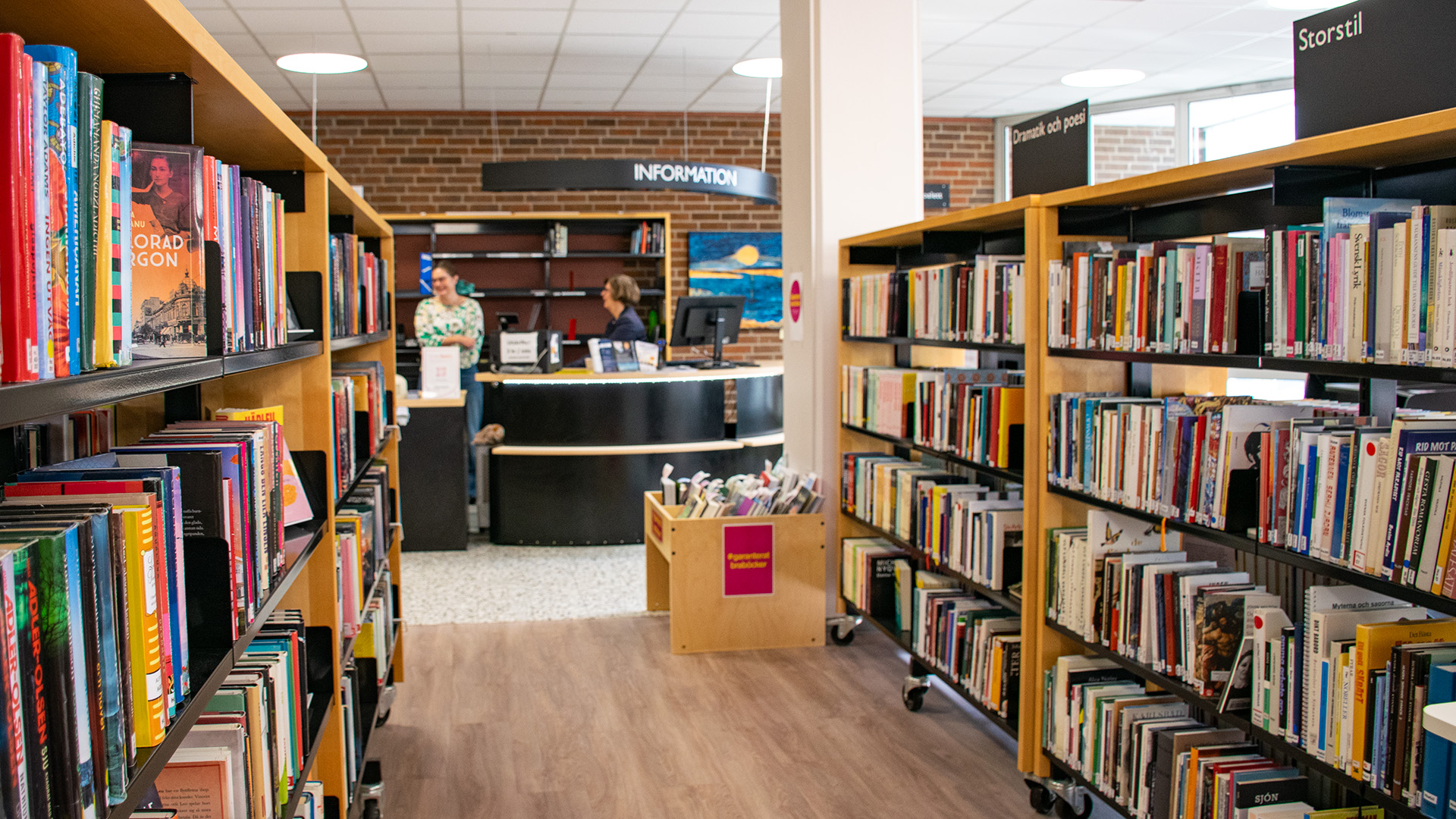 Bokhyllor fyllda med böcker står på varsin sida, längst fram i mitten finns bibliotekets informationsdisk. Två personer står bakom disken och pratar med varandra.