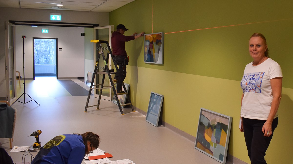 Konstnären Malin Palm står i en korridor där hennes två medhjälpare håller på att montera hennes målningar.