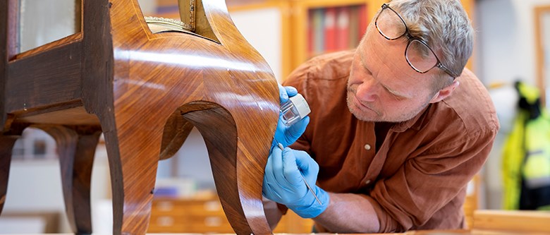 En man arbetar med ett litet verktyg och en lupp på ett träföremål. Han har ljusblå latexhandskar och glasögonen uppskjutna i pannan.