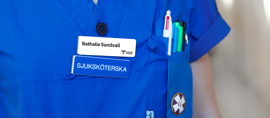 Två skyltar fäst på blå vårdkläder. En namnskylt med svart text och VGR:s logotyp på vit bakgrund. En yrkesskylt med titeln SJUKSKÖTERSKA i vit textfärg på blå bakgrund.