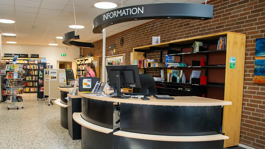 Informationsdisken på stadsbiblioteket i Strömstad är välvd och rundad i svart och ljust trä. På disken står skärmar och skyltar. Väggen bakom är i rött tegel liksom utsidan på huset.