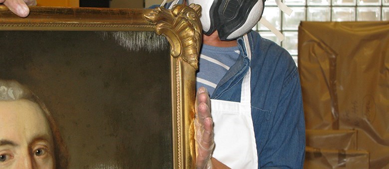 En man med ansiktsmask står och håller upp en ålderdomlig oljemålning med tydliga mögelfläckar. Mannen har latexhandskar på händerna.