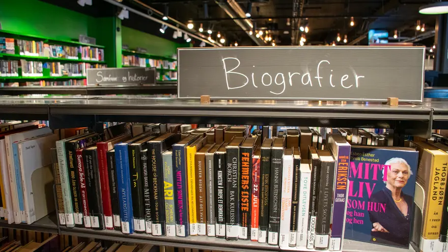 En låg bokhylla fylld med böcker. Ovanpå hyllan finns en skylt med texten "Biografier". Skylten är en svart tavla och texten skriven med vit krita.
