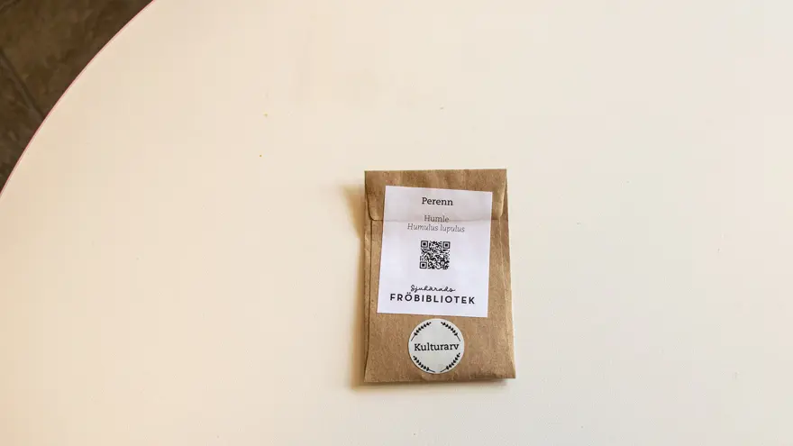 En brun liten fröpåse ligger på ett vitt bord. På den står det bland annat Humle, Perenn och Sjuhärads fröbibliotek. En QR-kod finns också på fröpåsen.