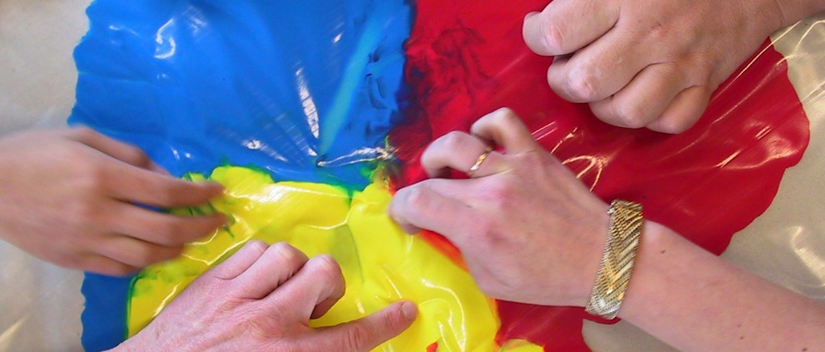 Flera händer trycker på plast som täcker över gul, röd och blå färg