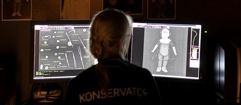 En kvinna sitter i ett mörkt rum framför två datorskärmar. På skärmarna syns röntgenbilder. Kvinnans tröja har texten "Konservator" tryckt på ryggen.