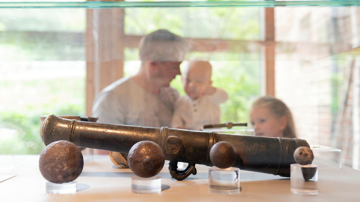En förälder med två småbarn bakom en glasmonter som innehåller en gammal metallkikare och kulor.