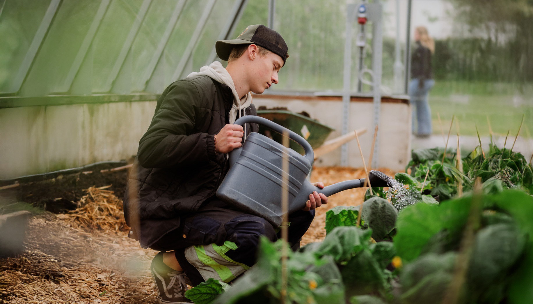 Elev sitter ned i växthus och håller i vattenkanna
